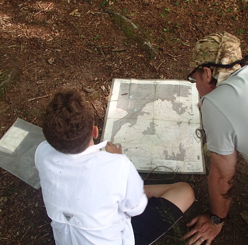 In the image, two individuals are examining a large map spread out on the ground. A younger person, possibly a child, is seated and appears to be pointing at something on the map with a pen or similar object. An older person, likely an adult, is leaning over the map, looking at it closely. The setting appears to be outdoors, possibly in a wooded area, as suggested by the ground covered with leaves and twigs.